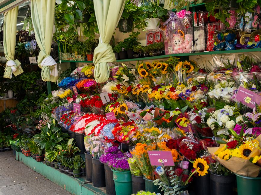 A vibrant floral display outside a flower shop showcases a variety of fresh flowers arranged in bouquets and potted plants. Prominent flowers include bright yellow sunflowers, white lilies, red roses,