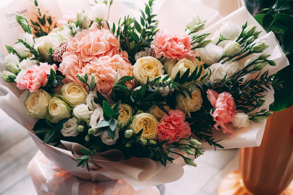 A bouquet of fresh flowers featuring soft pink carnations, creamy white roses, and pale yellow roses, accented with dark green foliage and sprigs of fern, wrapped in elegant cream-colored paper with a